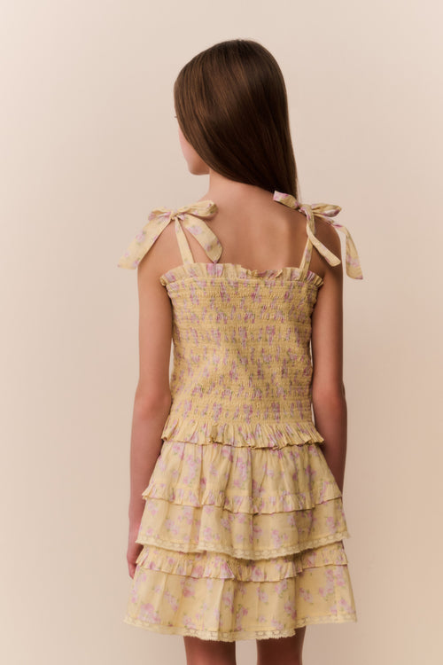 Tween girl wearing a yellow floral ruffle top and matching skirt with tiered layers on a beige background