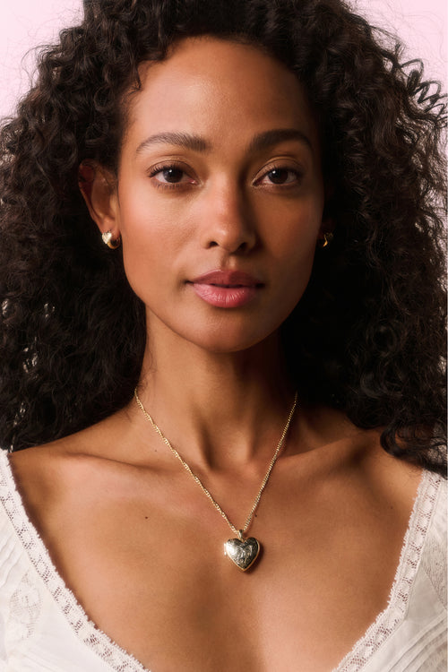 Woman wearing a gold necklace with a heart pendant against a pink background