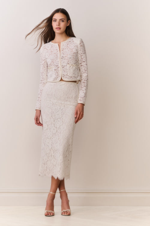Woman wearing a white lace jacket and skirt outfit on a plain background