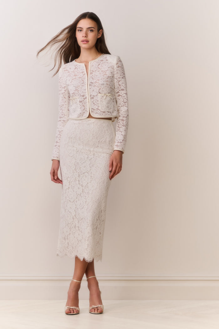 Woman wearing a white lace jacket and skirt outfit on a plain background