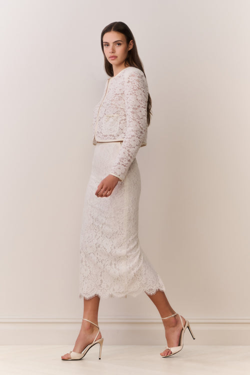 Woman wearing a white lace jacket and skirt outfit on a plain background