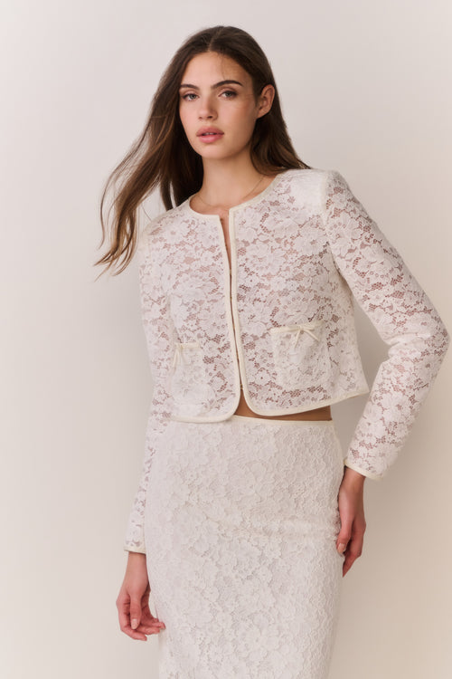 Woman wearing a white lace jacket and skirt outfit on a plain background