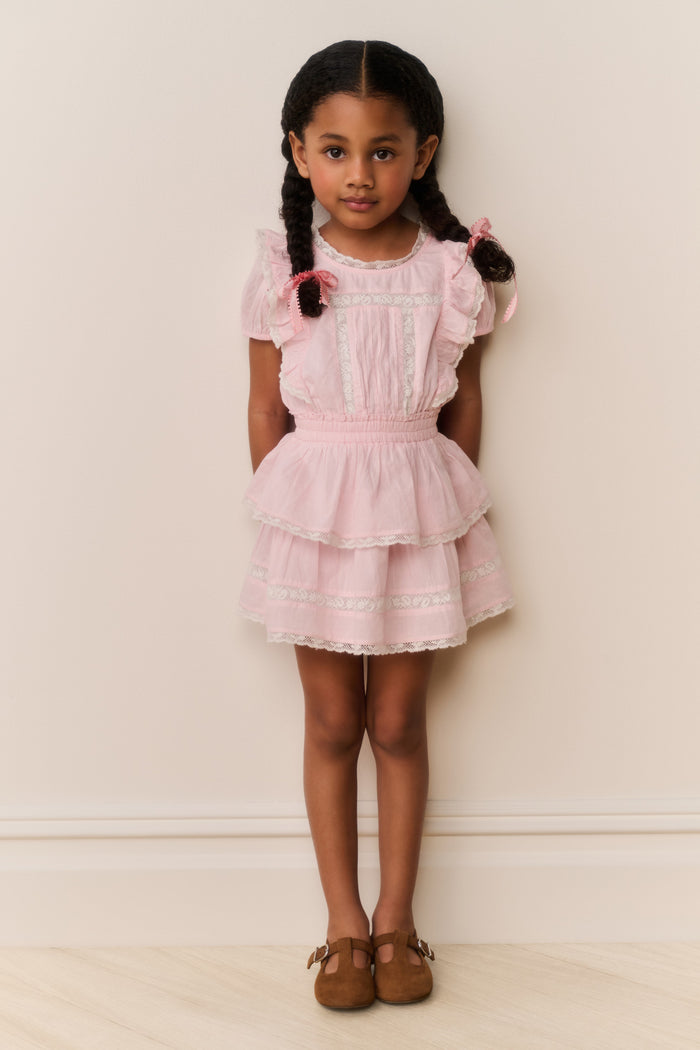 Young girl wearing a pink dress with ruffled sleeves and layers, standing against a plain background.