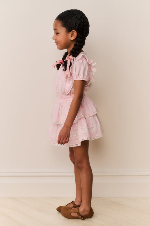 Young girl wearing a pink dress with ruffled sleeves and layers, standing against a plain background.