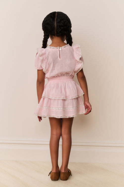 Young girl wearing a pink dress with ruffled sleeves and layers, standing against a plain background.
