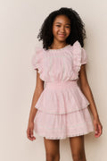 Young girl wearing a pink dress with ruffled sleeves and layers on a plain background