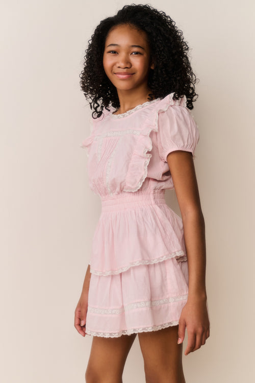 Young girl wearing a pink dress with ruffled sleeves and layers on a plain background