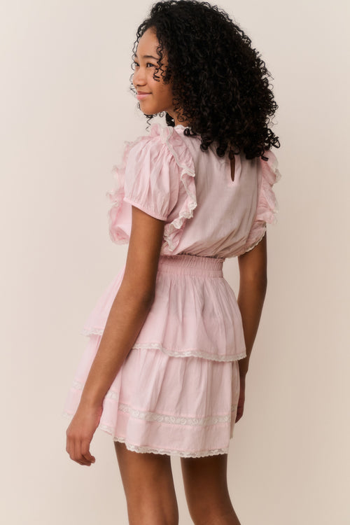 Young girl wearing a pink dress with ruffled sleeves and layers on a plain background