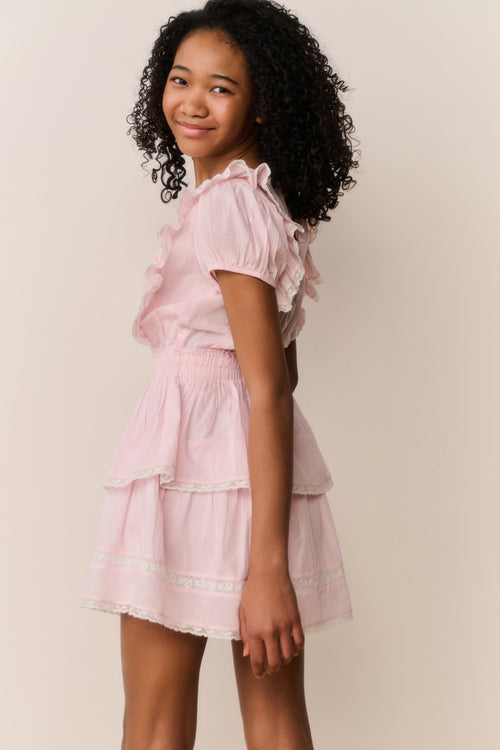 Young girl wearing a pink dress with ruffled sleeves and layers on a plain background