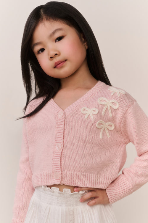 Young girl wearing a pink cardigan with bow details and white lace skirt against a plain background