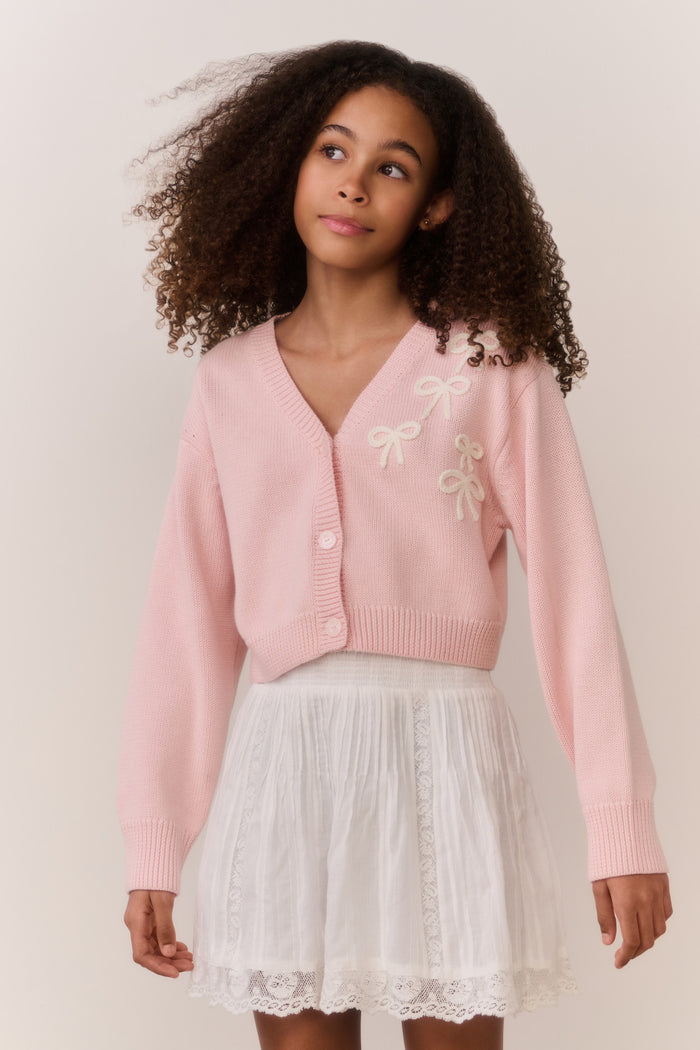 Tween wearing a pink sweater and white lace skirt on a plain background