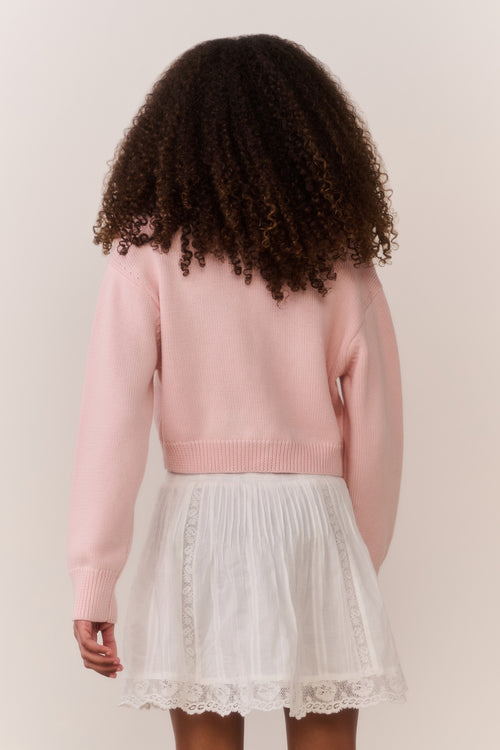 Tween wearing a pink sweater and white lace skirt on a plain background