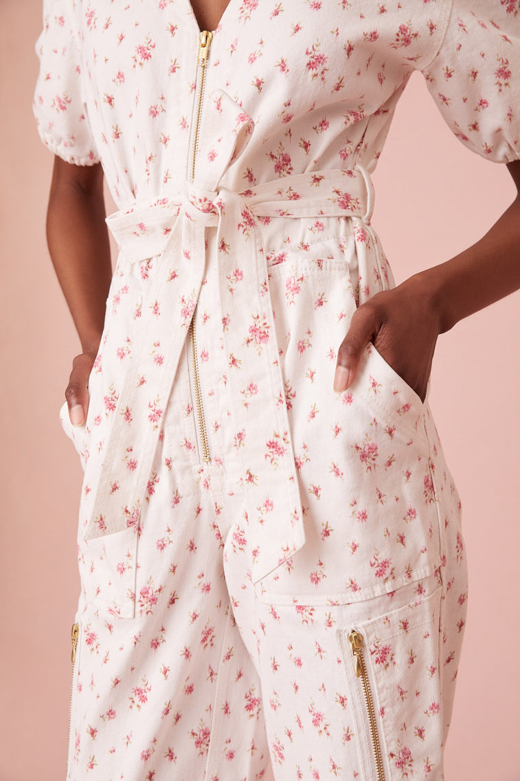 Jumpsuit featureing a slightly faded floral print on a stone-washed denim fabric. Features short puff sleeves, a v-neckline, cinching at the waist, elastic at the back, and a midi length. Includes a zipper at the front and a snap at the leg.
