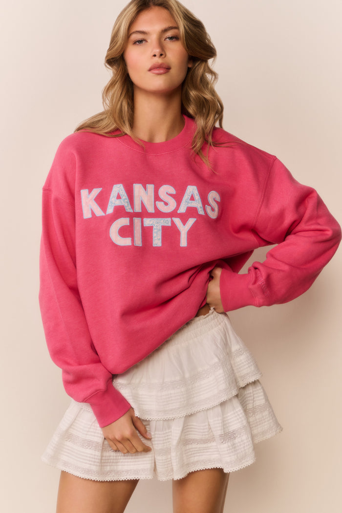 Woman wearing a pink 'Kansas City' sweatshirt with a white ruffle skirt and a beige background