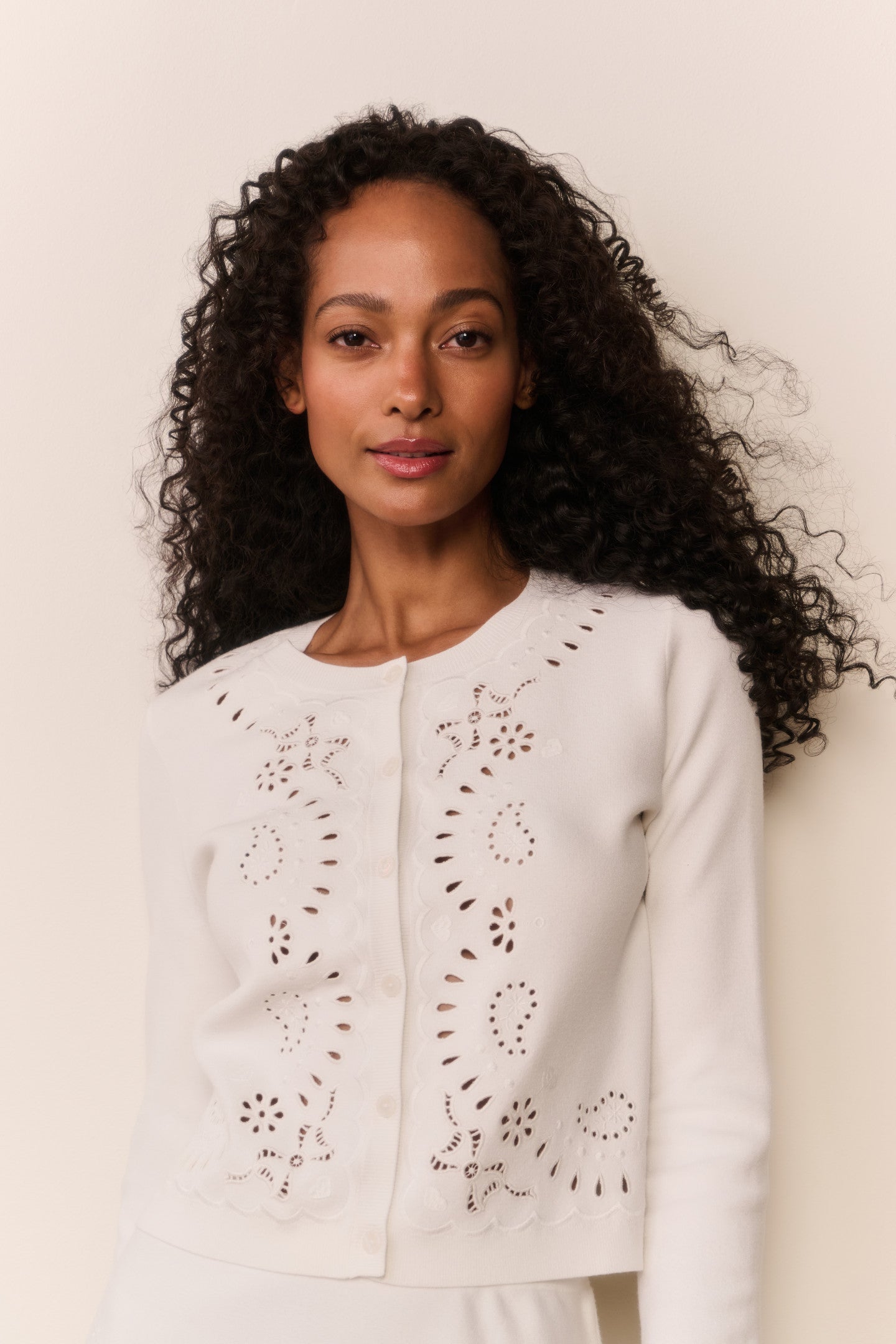 Woman wearing a white cardigan with intricate eyelet patterns on a plain background