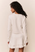 Woman wearing a white cardigan with intricate eyelet patterns and matching skirt on a plain background