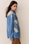 Woman wearing Blue denim shirt with floral lace embroidery and suede brown pants on a beige background