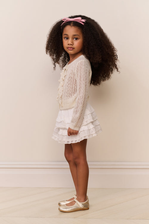 Young girl in a white dress and cream cardigan standing against a plain background
