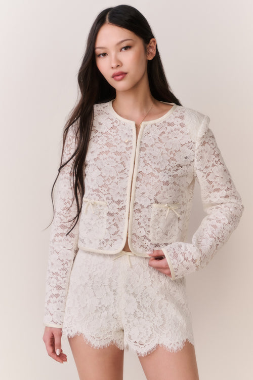 Woman wearing a white lace jacket and shorts outfit on a plain background