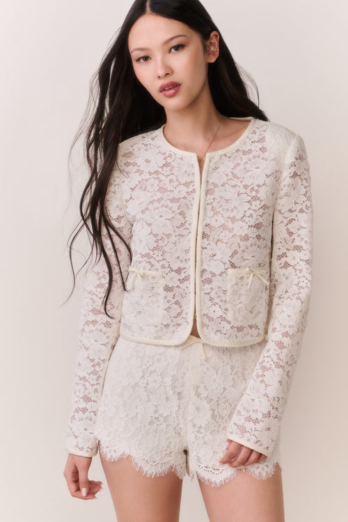 Woman wearing a white lace jacket and shorts outfit on a plain background