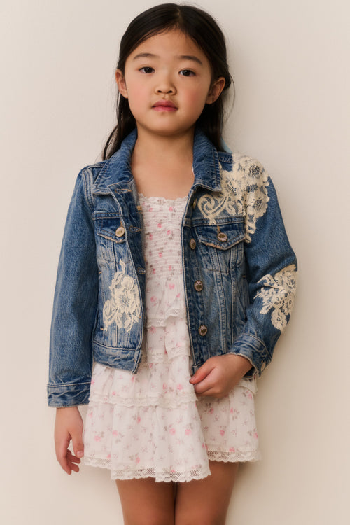 Young girl wearing a denim jacket with white floral lace embroidery on a white background with a floral skirt and a blue ribbon in their hair against a beige background