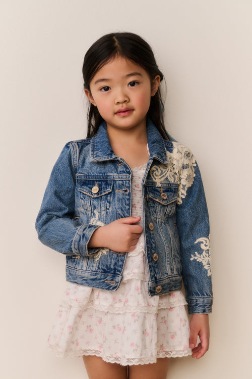 Young girl wearing a denim jacket with white floral lace embroidery on a white background with a floral skirt and a blue ribbon in their hair against a beige background