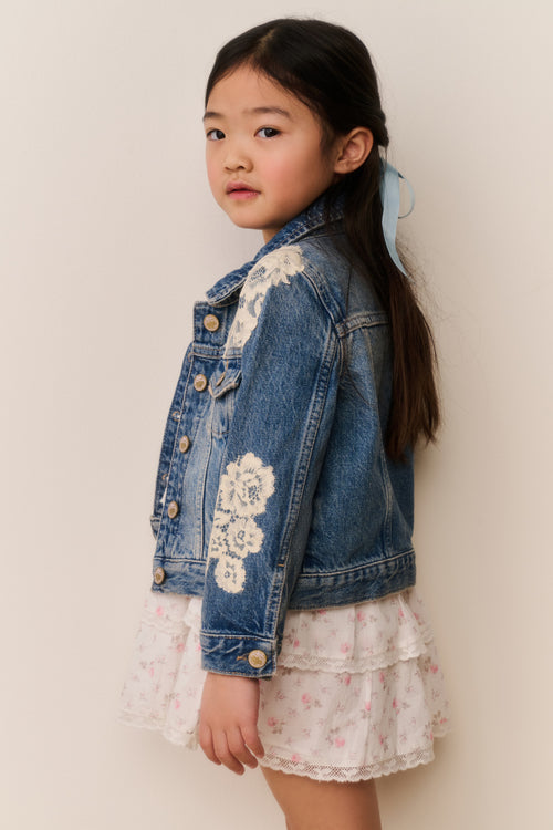 Young girl wearing a denim jacket with white floral lace embroidery on a white background with a floral skirt and a blue ribbon in their hair against a beige background