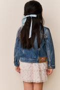 Young girl wearing a denim jacket with white floral lace embroidery on a white background with a floral skirt and a blue ribbon in their hair against a beige background