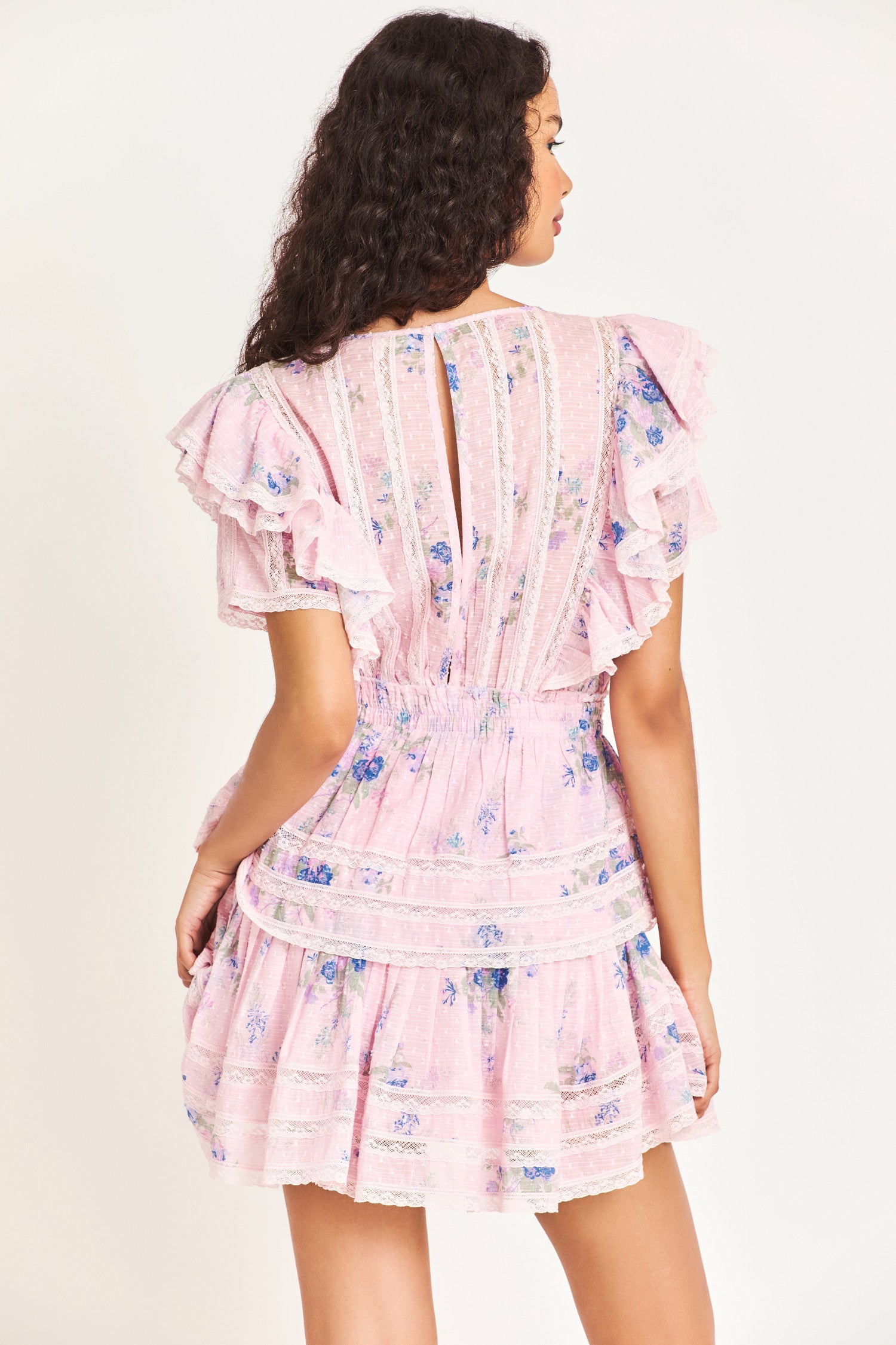The Natasha dress in pink features double ruffled flutter sleeves, an elasticated waist, and custom lace panels at the trim of the skirt. The dress also features small blue flower prints throughout the dress. 