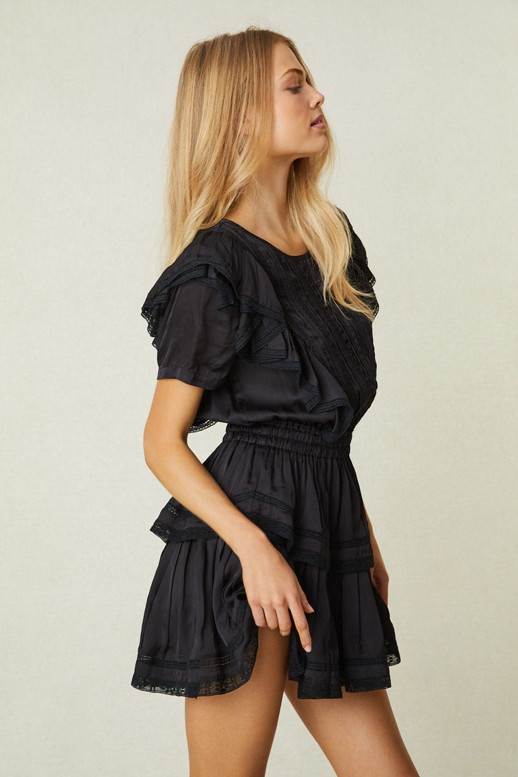Side image of model wearing black mini dress with ruffled skirt and shoulders and lace detail
