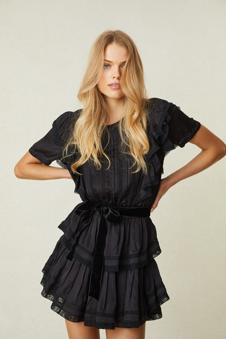 Model wearing black mini dress with ruffled skirt and shoulders and lace detail