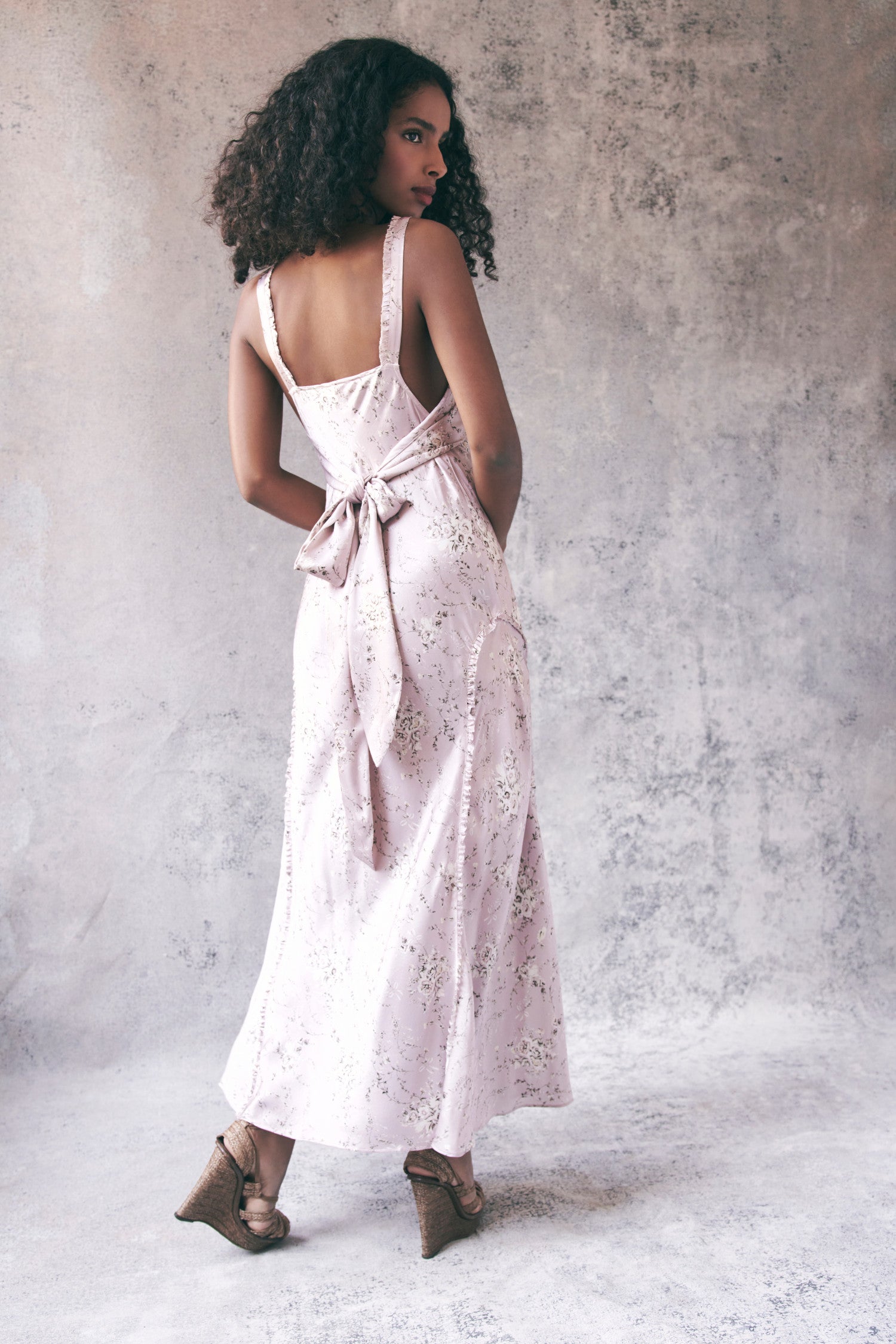 Back image of model wearing light pink floral maxi dress with ruffle trim detail. Dress ties in back with a bow