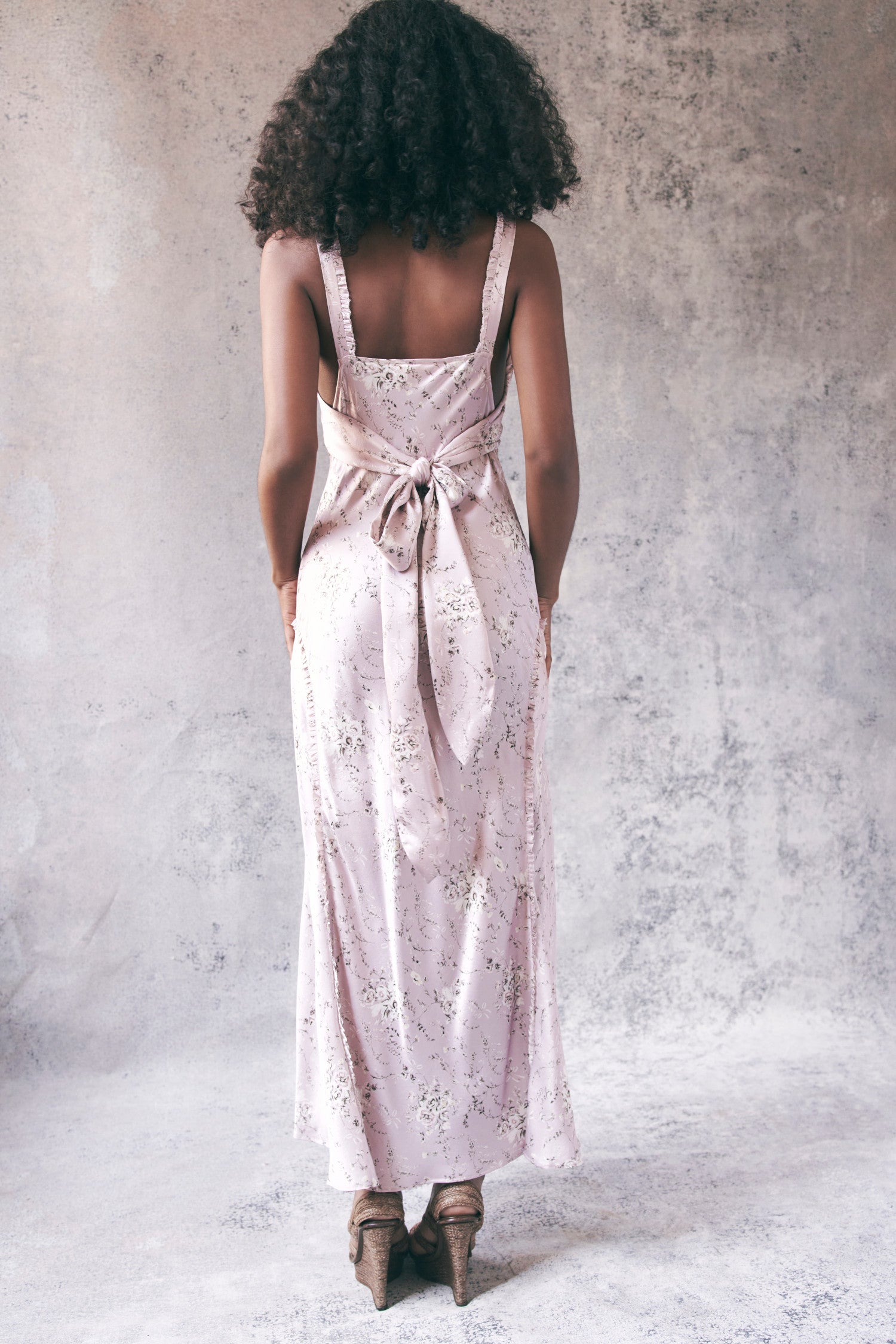 Back image of model wearing light pink floral maxi dress with ruffle trim detail and bow to tie on back