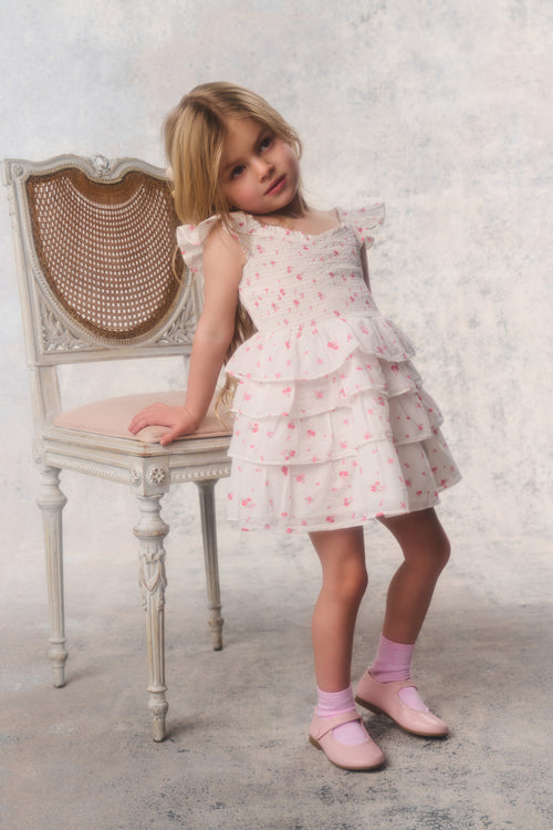 Young girl in a floral dress standing next to a chair against a textured white background