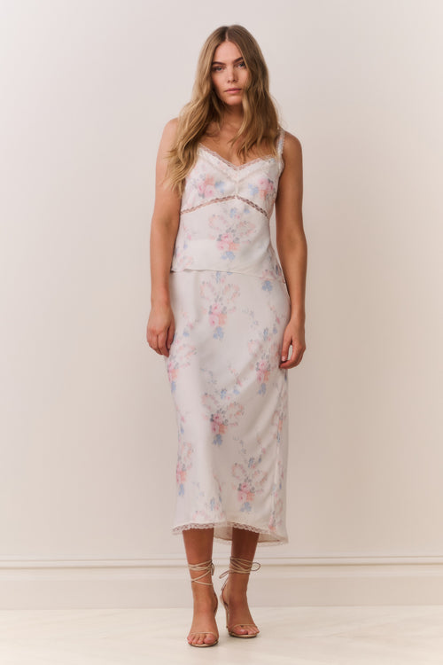 Woman wearing a floral satin lace trim camisole with thin straps and matching skirt on a plain background