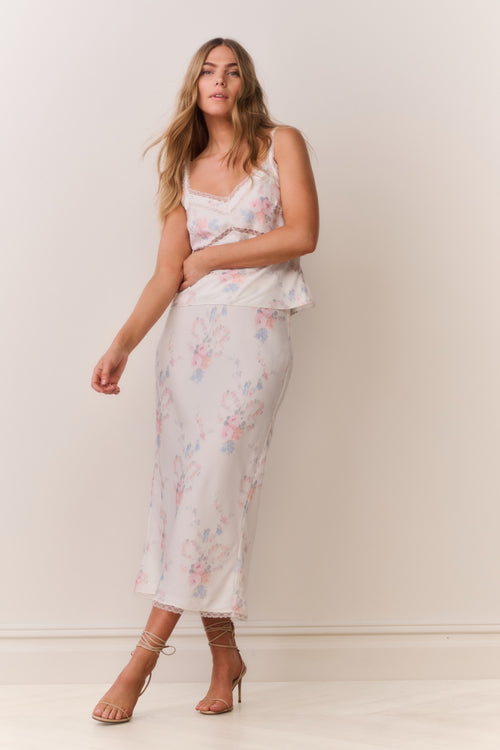 Woman wearing a floral satin lace trim camisole with thin straps and matching skirt on a plain background