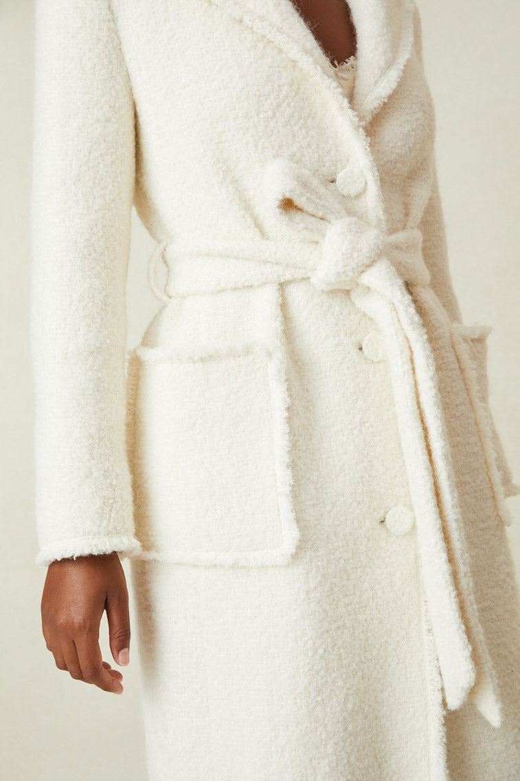 Close up image detailing fabric, button up closure, waist tie, and pockets on white midi length coat. 