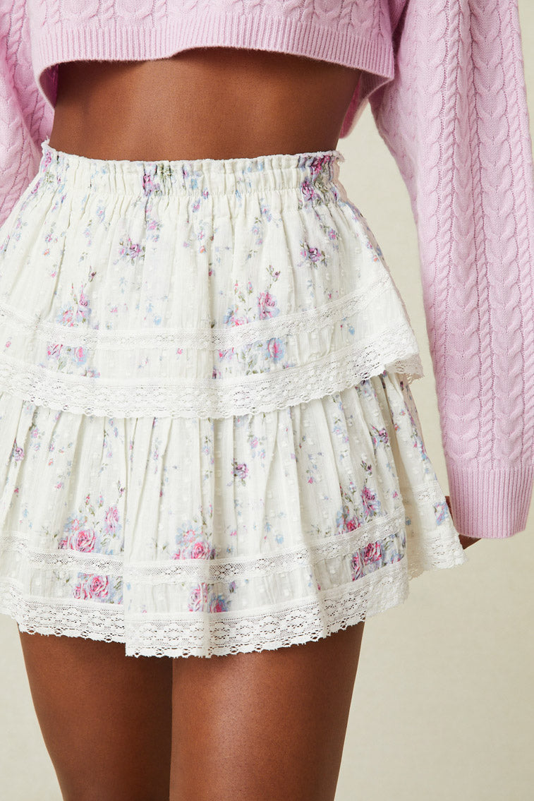 Model wearing white ruffle mini skirt with pink and purple floral print. Shows lace detail at trim of ruffles. 