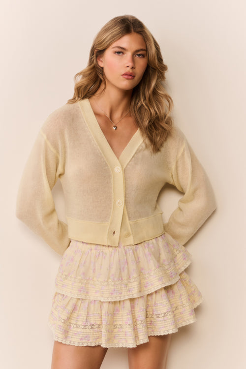 Woman wearing a pale yellow-colored cardigan over a patterned ruffle skirt on a plain background