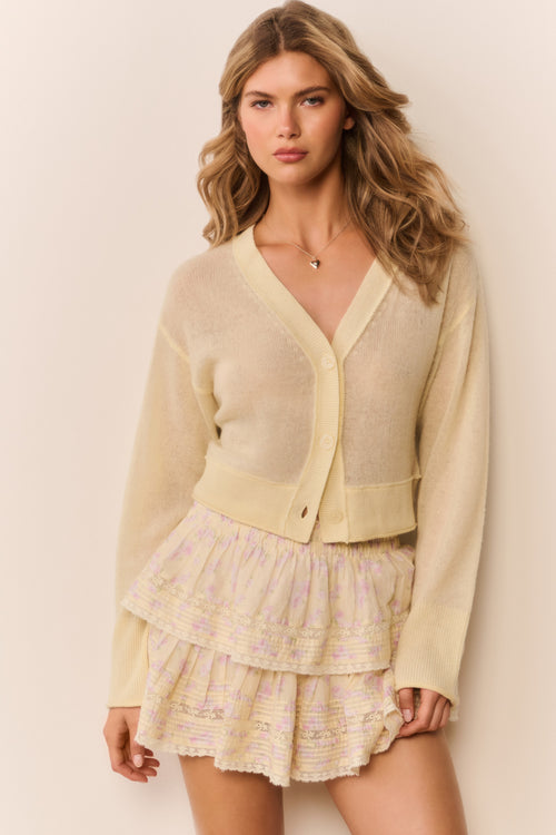 Woman wearing a pale yellow-colored cardigan over a patterned ruffle skirt on a plain background