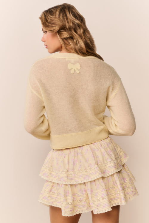 Woman wearing a pale yellow-colored cardigan over a patterned ruffle skirt on a plain background
