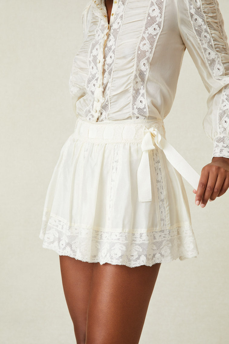 Model wearing white mini skirt with lace detailing at hem and ribbon with tie at waist. 