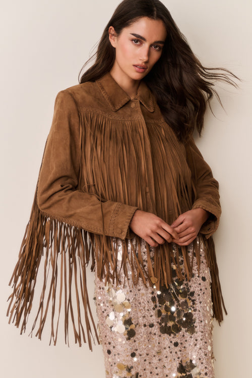 Woman wearing a brown suede fringed jacket and gold sequin dress with a plain background