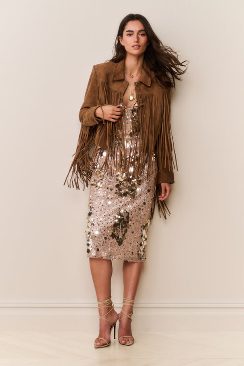Woman wearing a brown suede fringed jacket and gold sequin dress with a plain background