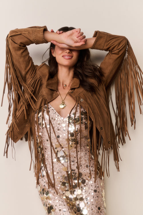 Woman wearing a brown suede fringed jacket and gold sequin dress with a plain background