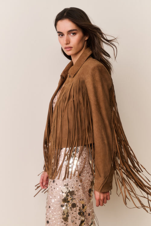 Woman wearing a brown suede fringed jacket and gold sequin dress with a plain background
