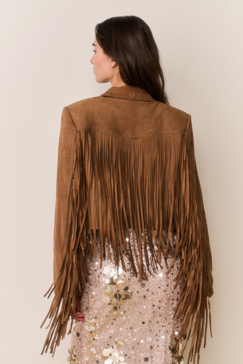 Woman wearing a brown suede fringed jacket and gold sequin dress with a plain background