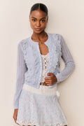 Woman wearing a light blue ruffle lace cardigan over a Skirt and top with blue floral pattern and white lace trim with a beige background