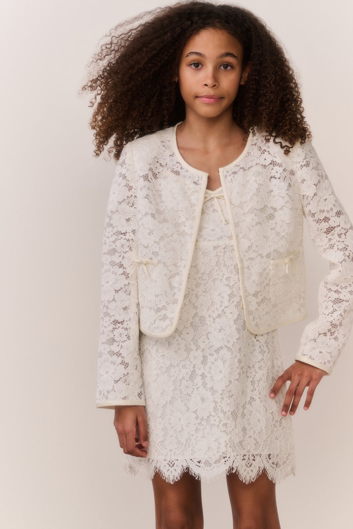 Tween wearing a white lace jacket and mini dress outfit on a plain background