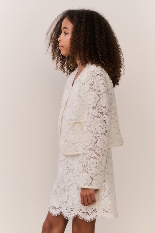 Tween wearing a white lace jacket and mini dress outfit on a plain background